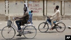 Dua remaja naik sepeda di kota Kano, Nigeria dengan tembok bertuliskan "Stop Membunuh Muslim, Tentara". Tentara Nigeria dituduh membantai Muslim Syiah di Kaduna dan menguburnya secara massal. 
