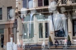 A woman sitting on a stoop reading a book in the sun is seen reflected by a closed clothing store's window on West Broadway on May 7, 2020, in the SoHo neighborhood of the Manhattan borough in New York.